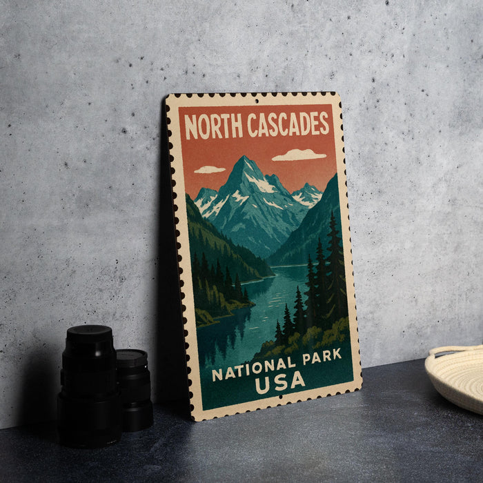 A vintage-style metal sign for North Cascades National Park in the United States, featuring a mountain landscape with a river and pine trees.