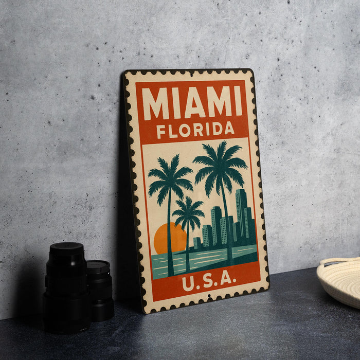 A vintage-style metal sign with the text "MIAMI FLORIDA" and a graphic of palm trees and a sunset, placed on a gray surface next to some black camera lenses.