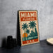 A vintage-style metal sign with the text "MIAMI FLORIDA" and a graphic of palm trees and a sunset, placed on a gray surface next to some black camera lenses.