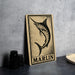 a wooden sign with a black and white illustration of a marlin fish, placed on a gray countertop next to some black camera lenses.