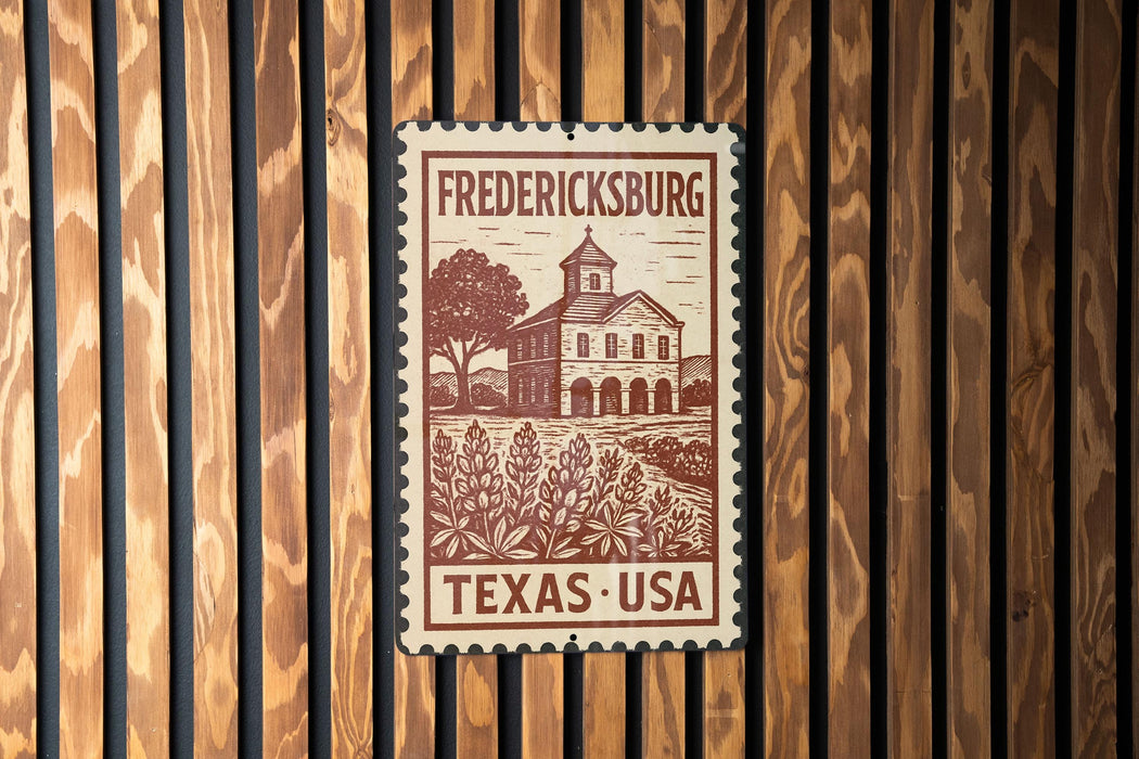 A metal sign with a wooden border displaying the text "FREDERICKSBURG TEXAS - USA" and an illustration of a building surrounded by trees.