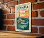 A vintage-style metal sign with the text "OLYMPIA WASHINGTON U.S.A." displayed prominently, featuring a mountain landscape and a building.