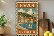 a vintage-style metal stamp with a depiction of a coastal town in Croatia, surrounded by a body of water and a sailboat. The stamp is placed on a wooden surface, with a potted plant to the left and a wooden cutting board to the right. There are also some lemons visible in the image.