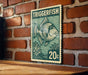 A green and white postage stamp featuring a fish illustration is displayed on a wooden surface against a brick wall.