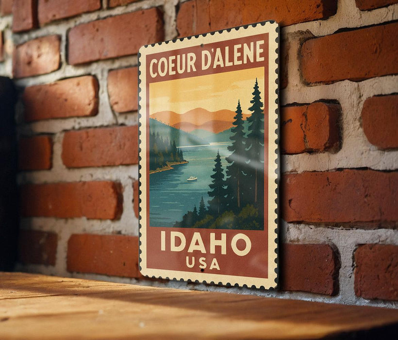 A vintage-style postage stamp depicting Coeur d'Alene, Idaho, USA, with a scenic landscape of mountains, pine trees, and a lake.