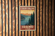 A vintage-style postage stamp from Idaho, USA, featuring a scenic landscape with pine trees, a lake, and mountains.