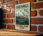 A vintage-style postage stamp featuring a mountain landscape and the text "DENALI NATIONAL PARK USA" is displayed on a brick wall.