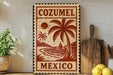 A vintage-style metal sign with the text "COZUMEL MEXICO" printed on it, featuring a palm tree and a sunset, is displayed on a shelf next to a wooden cutting board and a potted plant.