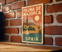 A vintage-style poster of the city of Majorca, Spain, featuring a castle and a sailboat, is displayed on a brick wall.