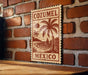 A wooden sign with the text "COZUMEL MEXICO" is displayed on a brick wall, featuring a palm tree and a sailboat illustration.