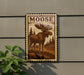 A metal plaque featuring a moose illustration is mounted on a wall, with a plant to the left.