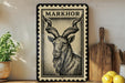 a framed illustration of a horned goat, with a wooden cutting board and a potted plant in the background.