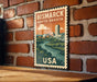 A vintage-style metal stamp depicting a cityscape with a river, mounted on a brick wall.