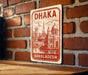 A sign displaying the name "Dhaka" and the word "Bhangladesh" is hanging on a brick wall.