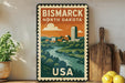 A vintage-style metal sign with a colorful illustration of a cityscape and a river, along with the text "BISMARK NORTH DAKOTA USA".