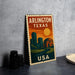 A vintage-style metal sign with the text "ARLINGTON TEXAS USA" printed on it, featuring an illustration of a city skyline at sunset.