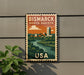 A vintage postage stamp depicting a cityscape with a river, mounted on a wall next to a plant.