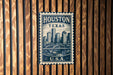 A vintage-style postage stamp from Texas, featuring a cityscape and a bridge, is displayed on a wooden wall.