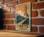 A vintage-style poster of a lighthouse and a coastal town, with the text "CRETE GREECE" prominently displayed, is hanging on a brick wall.