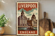 A vintage-style metal sign with the text "LIVERPOOL ENGLAND" and an illustration of a city skyline, including a clock tower, is displayed on a shelf next to a wooden cutting board and some potted plants.