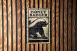 a metal sign with a picture of a badger and the text "HONEY BADGER" on it, mounted on a wooden wall.