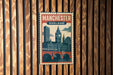 A vintage-style poster of Manchester, England, featuring a cityscape with a clock tower and a bridge, is displayed on a wooden wall.