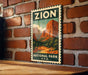 A vintage-style poster of Zion National Park, featuring a scenic mountain landscape, is displayed on a brick wall.