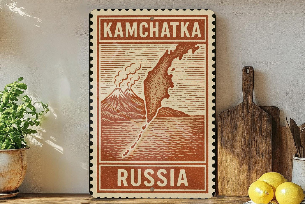 A vintage-style stamp or sign with the text "KAMCHATKA RUSSIA" printed on it, placed on a wooden surface next to a potted plant and some kitchen utensils.