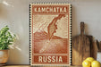 A vintage-style stamp or sign with the text "KAMCHATKA RUSSIA" printed on it, placed on a wooden surface next to a potted plant and some kitchen utensils.