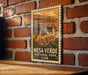 A framed poster of the mesa verde, a famous landmark in the National Park of the United States, is displayed on a brick wall.