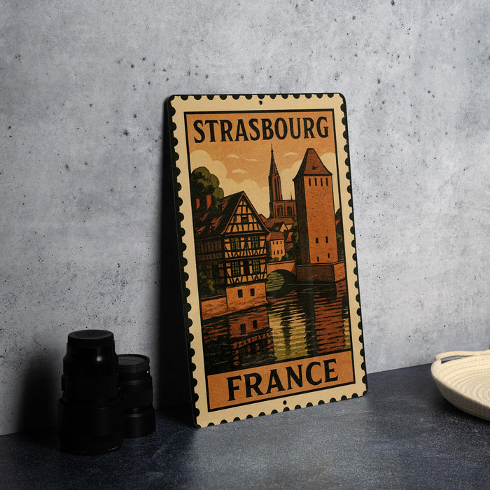 A vintage-style metal sign with a castle and buildings, featuring the text "STRASBOURG FRANCE" on it, is displayed on a table next to some black camera lenses.