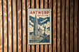 A vintage-style metal sign with the text "ANTWERP BELGIUM" displayed prominently, featuring an illustration of a clock tower and a statue.