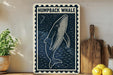 a framed artwork of a humpback whale on a wall, with a wooden cutting board and a potted plant to the side.