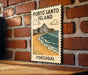 A vintage-style poster of Porto Santo Island, featuring a beach and ocean landscape, is displayed on a brick wall.