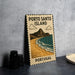 A vintage-style metal sign with the text "PORTO SANTO ISLAND" and an illustration of a beach and mountains, placed on a gray countertop next to some black camera lenses.