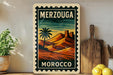 A vintage-style stamp depicting a desert landscape with a palm tree and mountains, with the text "MERZOUGA" and "MOROCCO" printed on it. The stamp is displayed on a shelf next to a wooden cutting board and a potted plant.