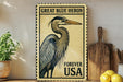 A vintage-style postage stamp featuring a great blue heron, with the text "GREAT BLUE HERON FOREVER USA" printed on it. The stamp is displayed on a shelf next to a wooden cutting board and a potted plant.