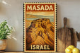 A framed vintage-style poster of Masada, a famous landmark in Israel, is displayed on a wall next to a wooden cutting board and some potted plants.