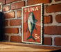 A vintage-style stamp featuring a fish, likely a tuna, is displayed on a brick wall.