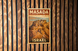 A vintage-style postage stamp depicting Masada, the ancient city of Jerusalem, is displayed on a wooden wall.