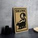a wooden sign with a black and white illustration of a skunk, placed on a gray countertop next to some black camera lenses.