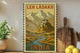 A framed vintage-style poster depicting a mountainous landscape with a small village or town in the center, surrounded by a winding river. The poster is titled "LEH LADAKH" and "INDIA", suggesting it is a travel poster for India. The poster is displayed on a wall next to a wooden cutting board and a potted plant, creating a cozy, homey atmosphere.