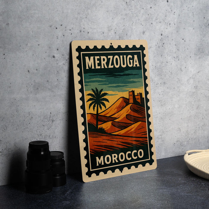 A vintage-style metal sign with a desert landscape and a palm tree, featuring the text "MERZOUGA MOROCCO" on it, is displayed on a table next to some black camera lenses.