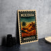 A vintage-style metal sign with a desert landscape and a palm tree, featuring the text "MERZOUGA MOROCCO" on it, is displayed on a table next to some black camera lenses.