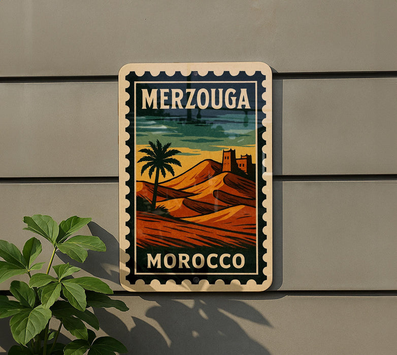 A vintage-style sign with the text "MERZOUGA" and "MOROCCO" printed on it, depicting a desert landscape with a palm tree and mountains. The sign is mounted on a wall, and there is a plant to the left of the sign.
