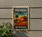 A vintage-style sign with the text "MERZOUGA" and "MOROCCO" printed on it, depicting a desert landscape with a palm tree and mountains. The sign is mounted on a wall, and there is a plant to the left of the sign.
