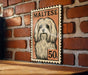 A framed illustration of a Maltese dog, with the text "MALTESE" and "50c" printed on it, is displayed on a brick wall.