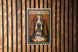 A metal sign with a picture of a Basset Hound on it, mounted on a wooden wall.