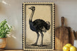 a framed illustration of an ostrich standing on a wooden surface, with a potted plant and a wooden cutting board visible in the background.