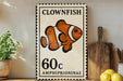 A vintage-style stamp featuring an illustration of a clownfish, with the text "CLOWNFISH" and "AMPHIPRIIONINAE" printed on it. The stamp is placed on a wooden surface, with a potted plant, a cutting board, and some lemons nearby.